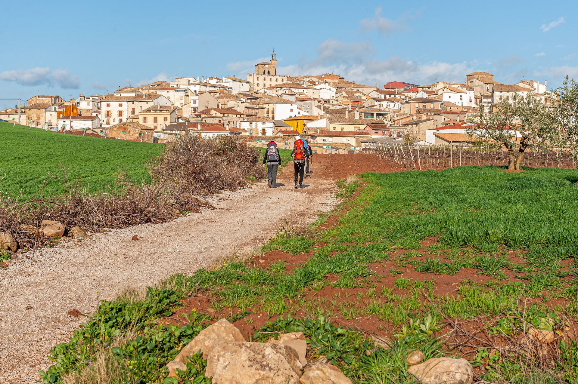 Dos excursionistas en un sendero de tierra a través de campos verdes, con un pueblo en la ladera de una colina al fondo.