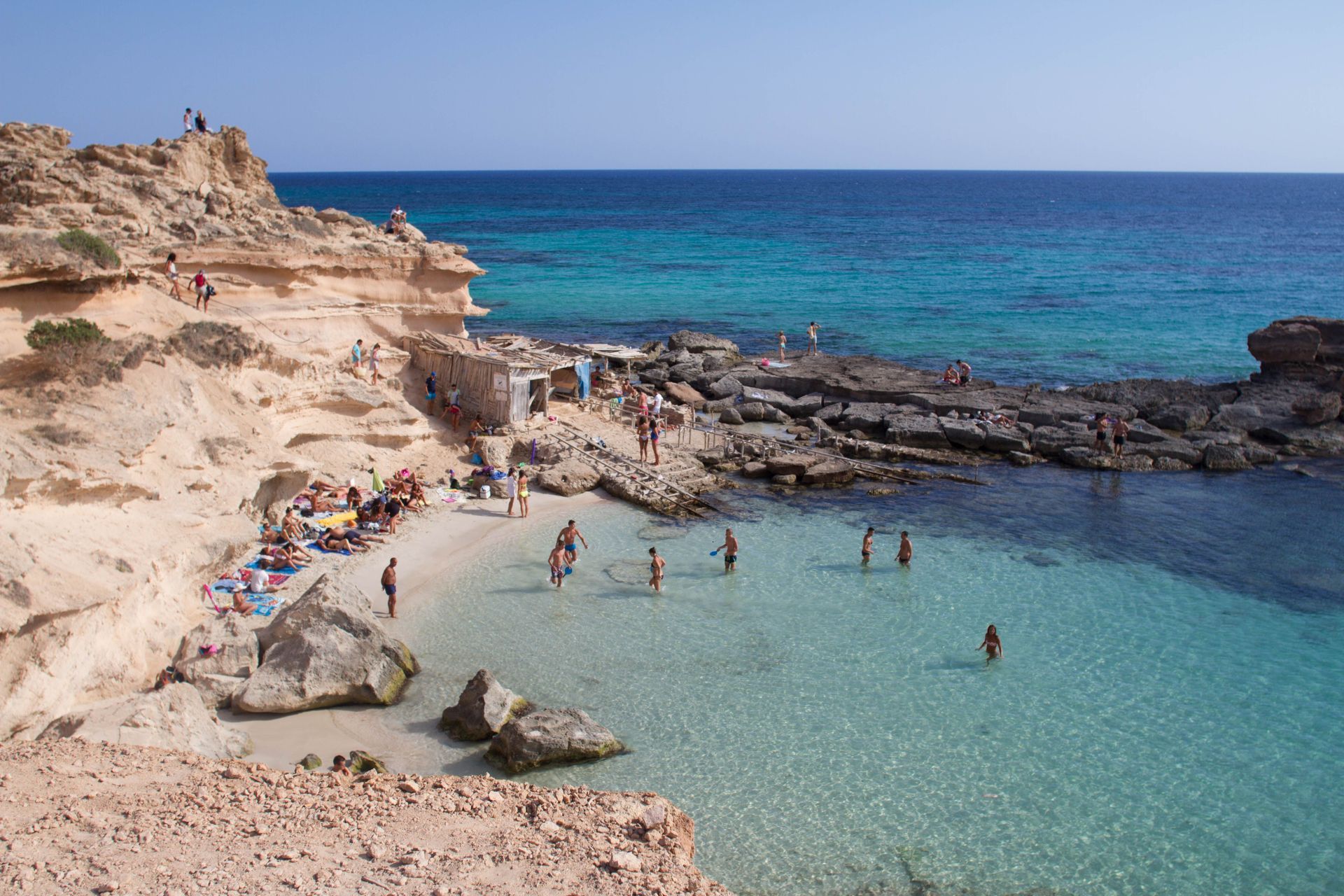 Gente nadando en una cala de aguas cristalinas de color turquesa, junto a acantilados rocosos y una playa de arena.