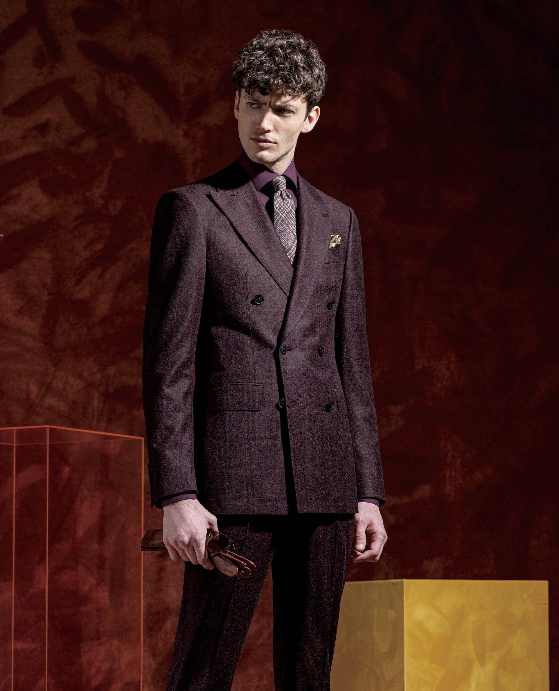 A man in a purple suit and tie is standing in front of a red wall.