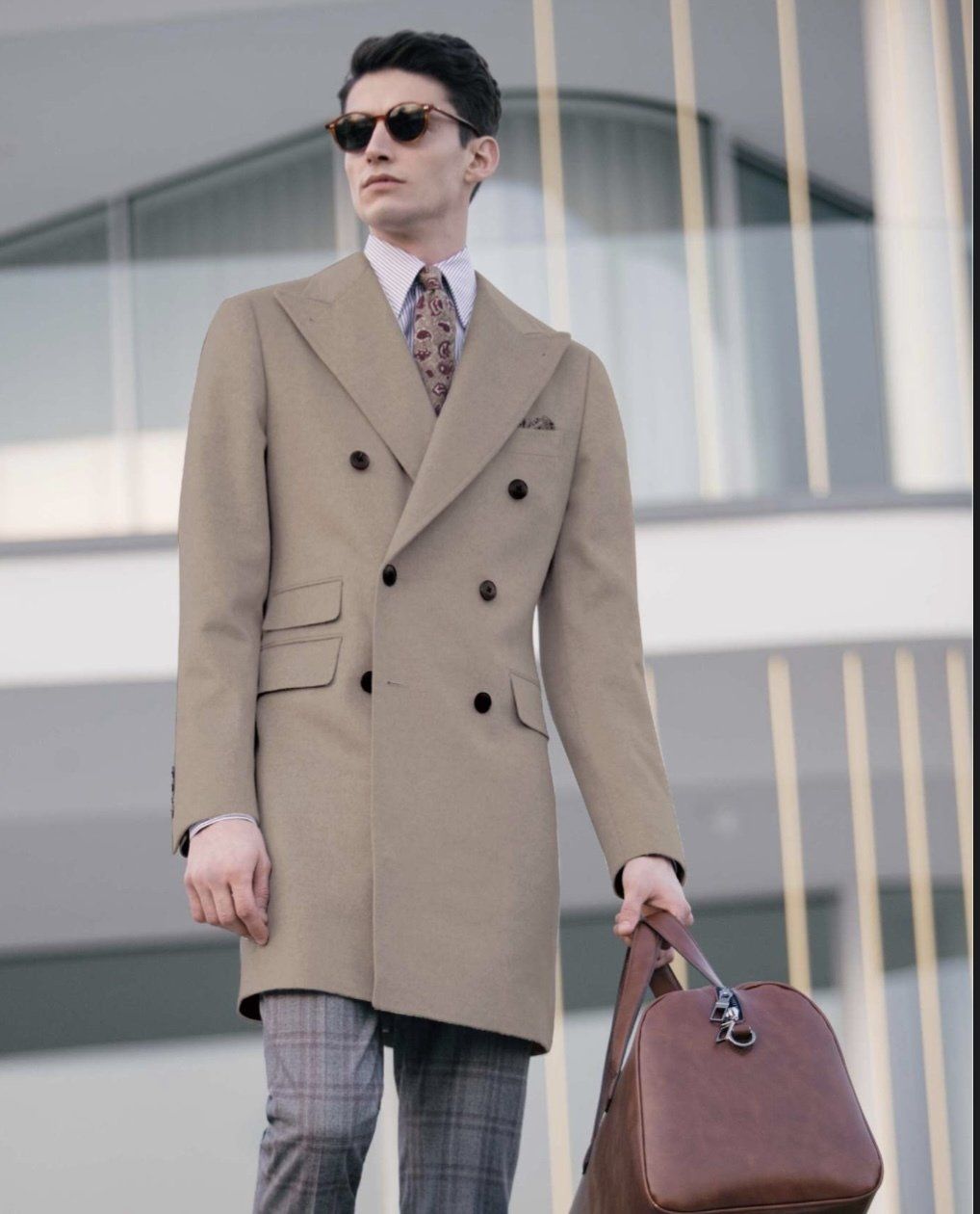 A man in a tan coat and tie is holding a brown bag