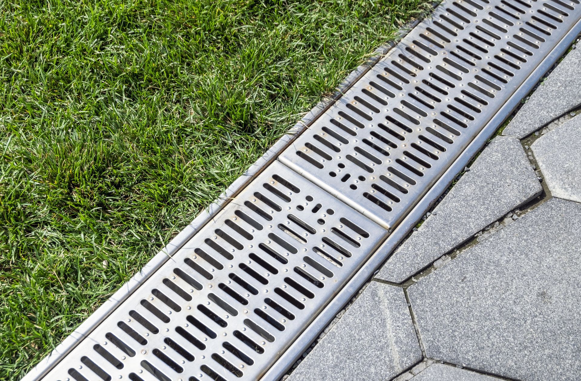 Metal drain along a paved walkway, next to a patch of green grass.