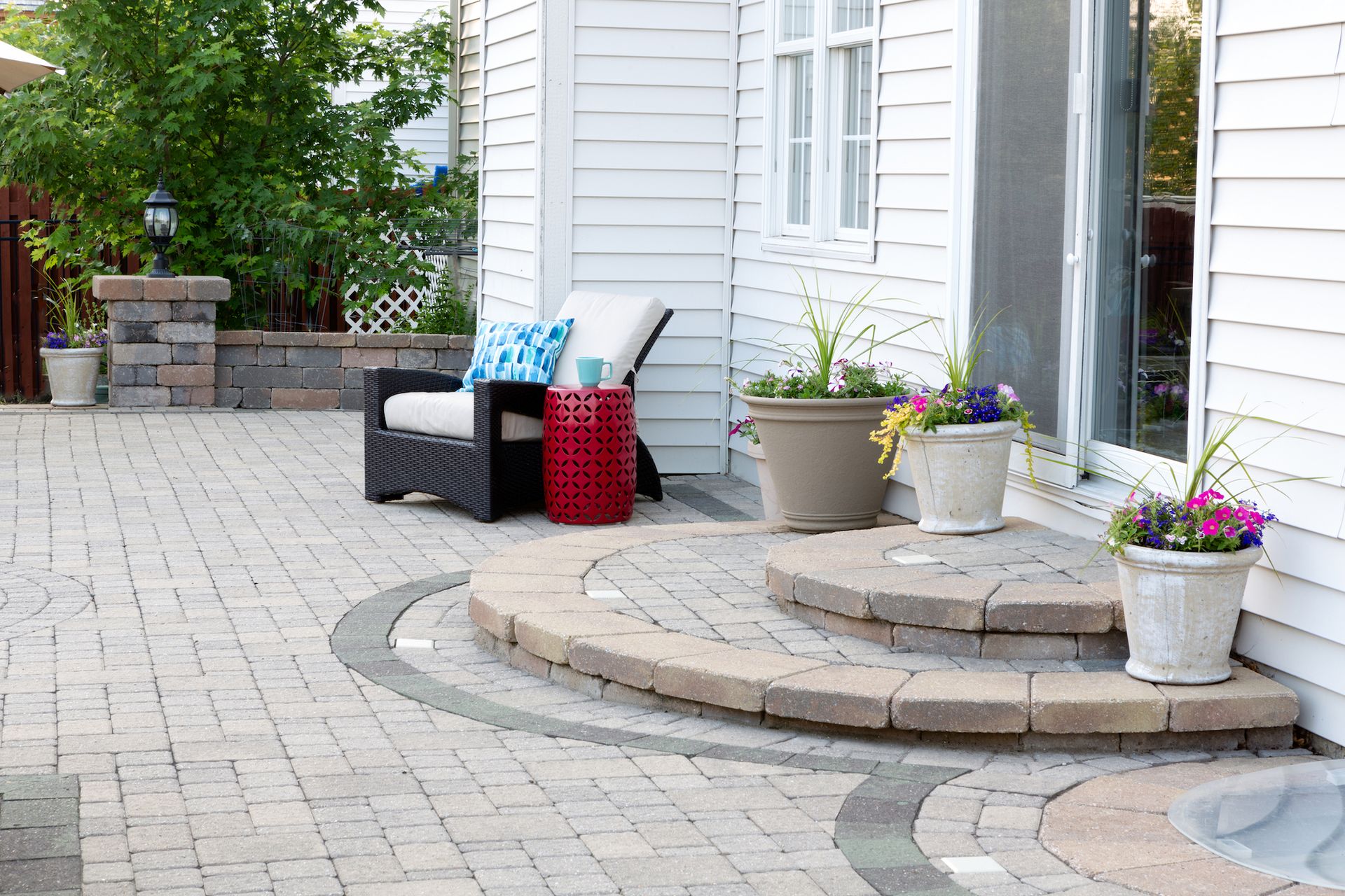 Stone patio with steps leading to a white house. Outdoor seating, potted flowers in view.