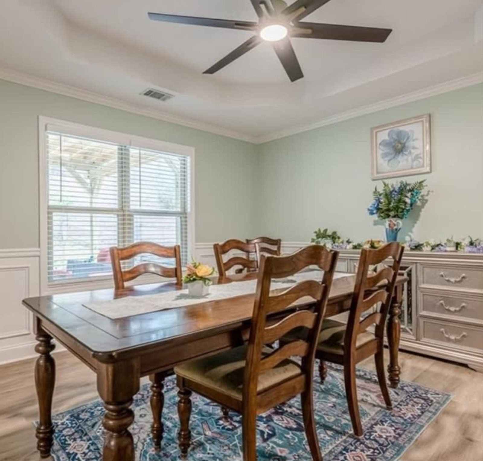 A dining room with a wooden table and chairs, a buffet, and a ceiling fan. The walls are light green, and there is a window.