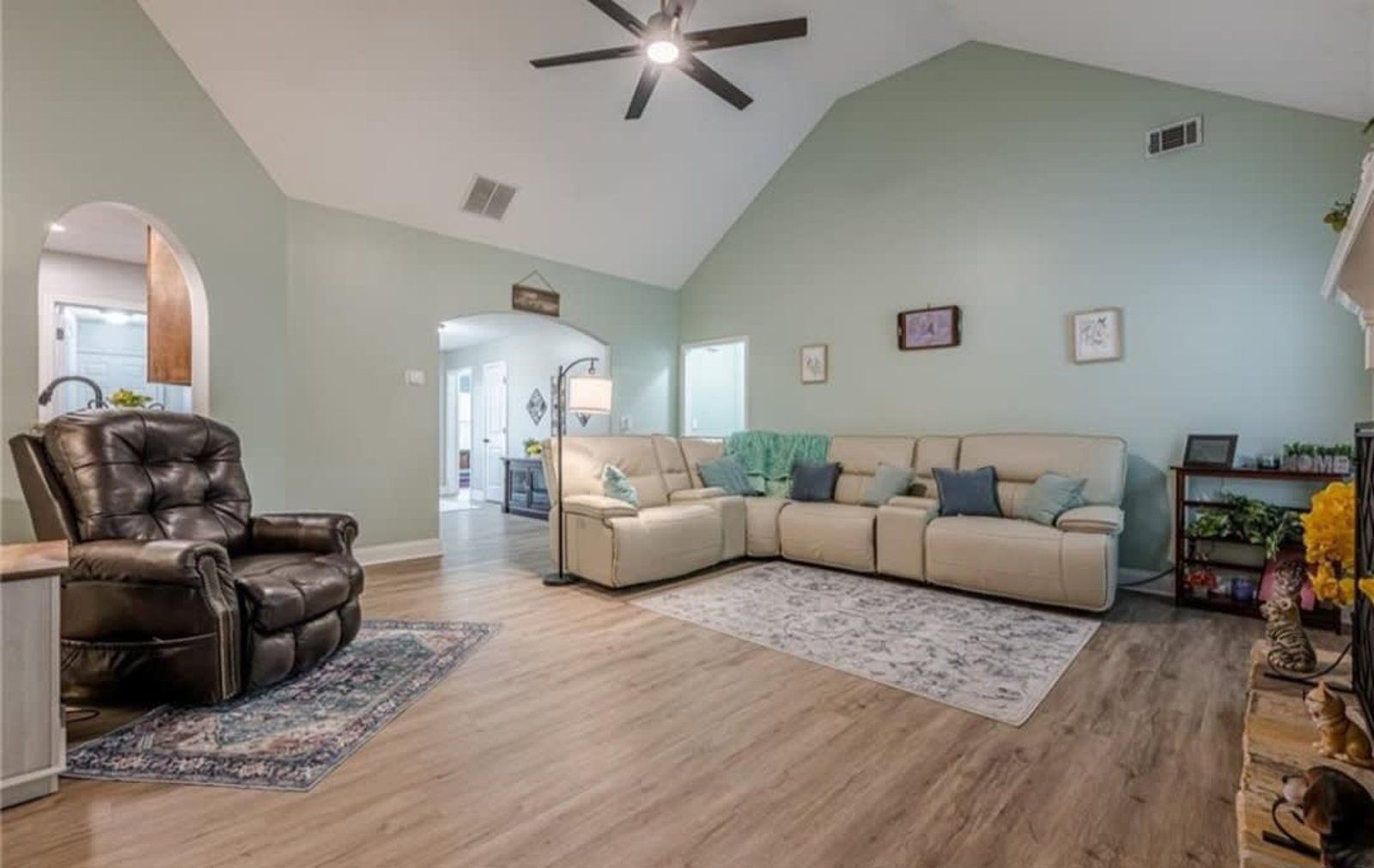 A living room with light green walls, a beige sectional sofa, a black leather recliner, and wood-look flooring.