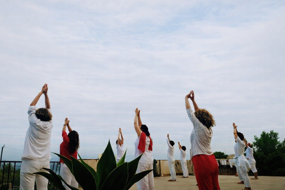 Persone vestite di bianco e rosso fanno yoga su un tetto con le mani alzate al cielo.