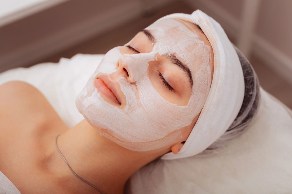 Donna con maschera facciale, occhi chiusi, appoggiata su una superficie ricoperta di bianco, in un ambiente termale.