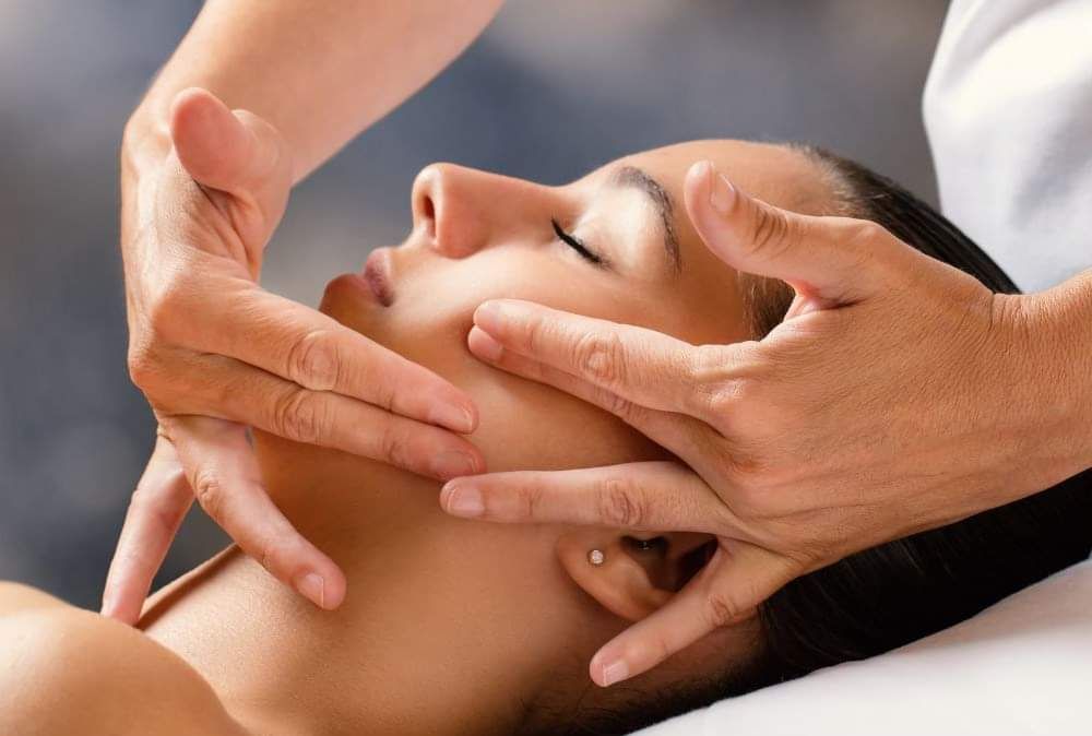 Mani che massaggiano il viso di una persona. Occhi chiusi. Sfondo blu e bianco.