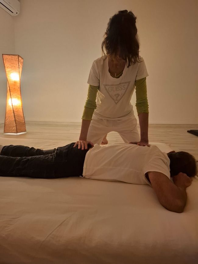 Persona che riceve un massaggio alla schiena su un lettino da massaggio; il terapista si inginocchia, esercitando pressione. Ambiente interno neutro.