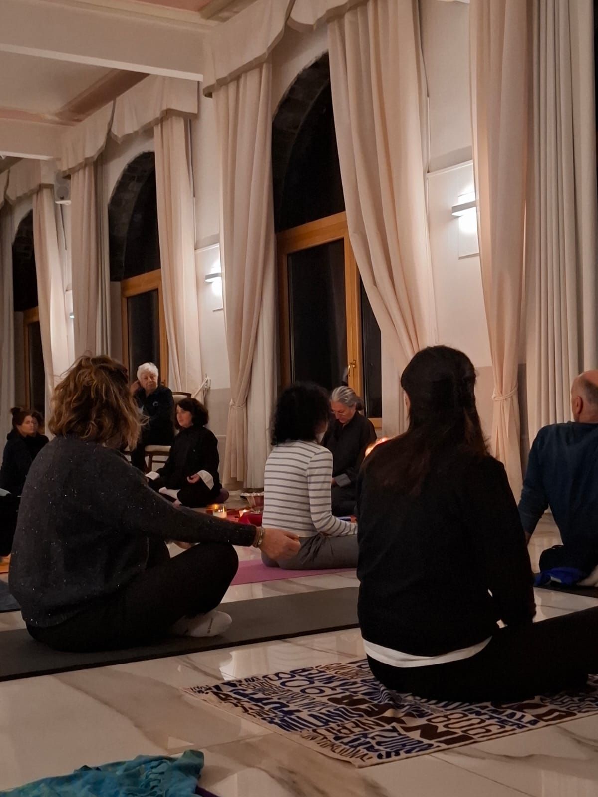 Persone che meditano in una stanza con alte finestre e tende. Sono sedute su stuoie. Illuminazione soffusa.