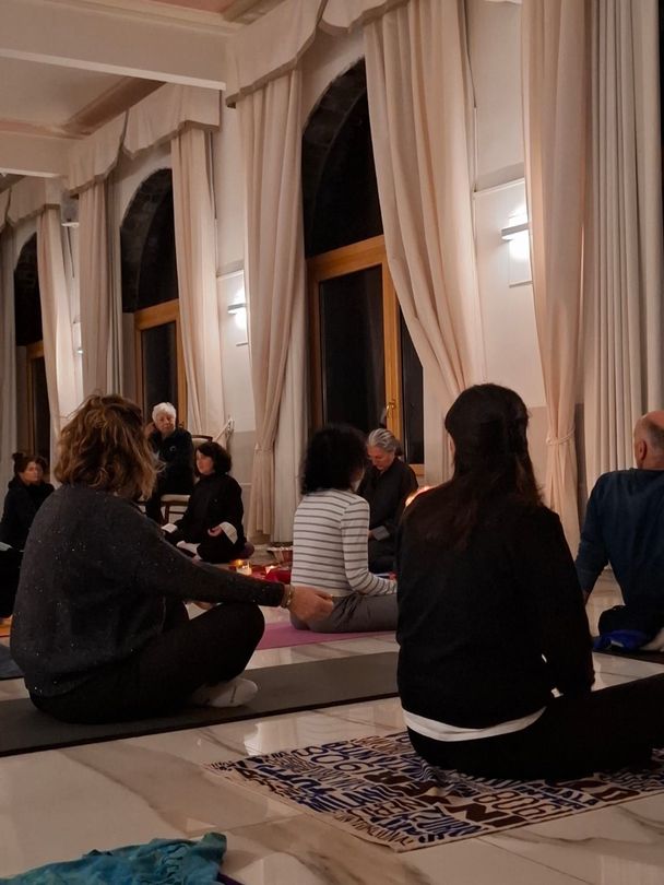 Persone che meditano in una stanza con alte finestre e tende. Sono sedute su stuoie. Illuminazione soffusa.