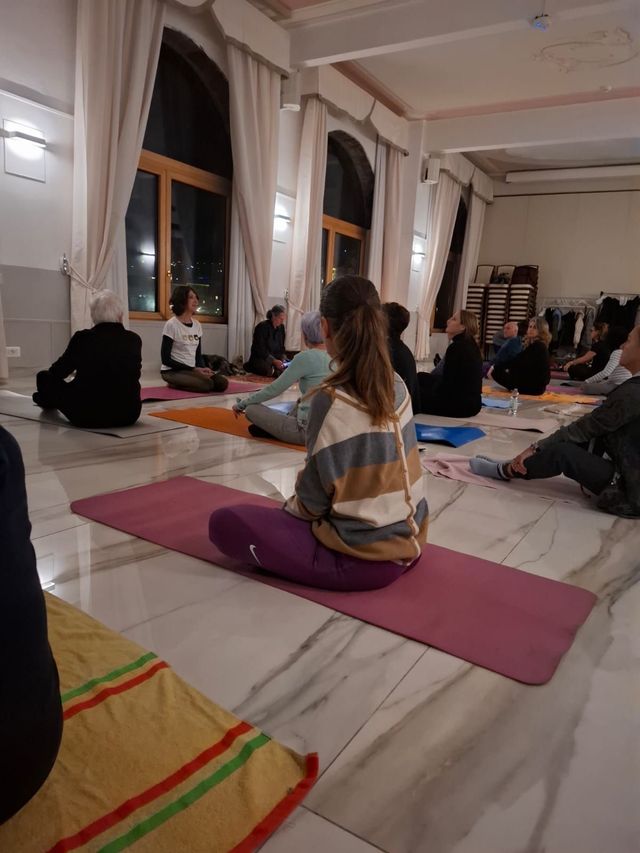 Persone sedute a gambe incrociate su tappetini da yoga in una stanza con grandi finestre, mentre si preparano per una sessione.