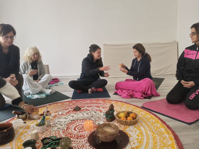 Le persone siedono in cerchio su stuoie, meditando e tenendo in mano oggetti. Al centro ci sono un tappeto mandala e delle candele.