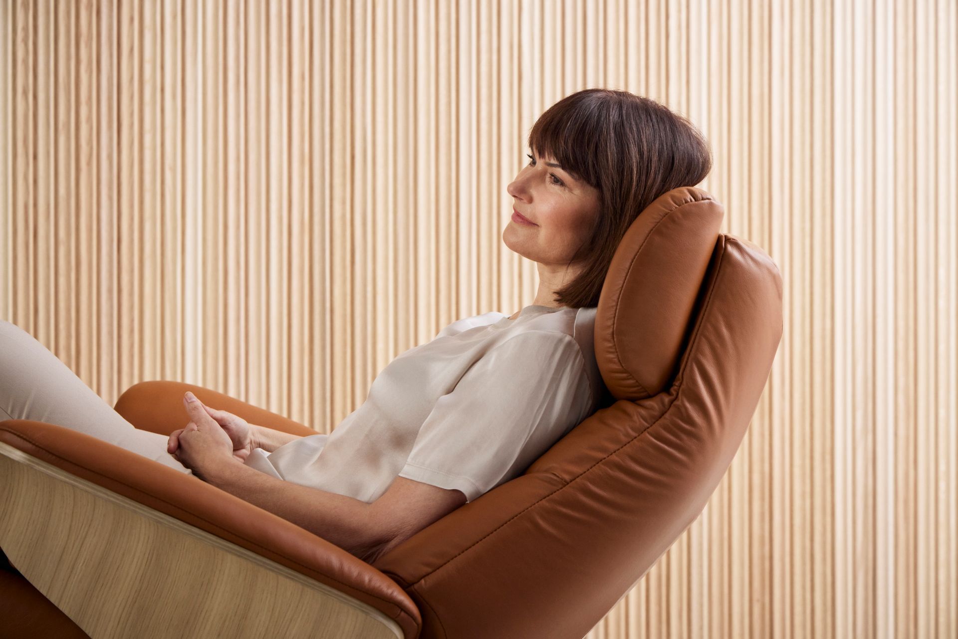 A person rests in a tan leather recliner against a vertical-slat wall, looking relaxed and glancing to the side.