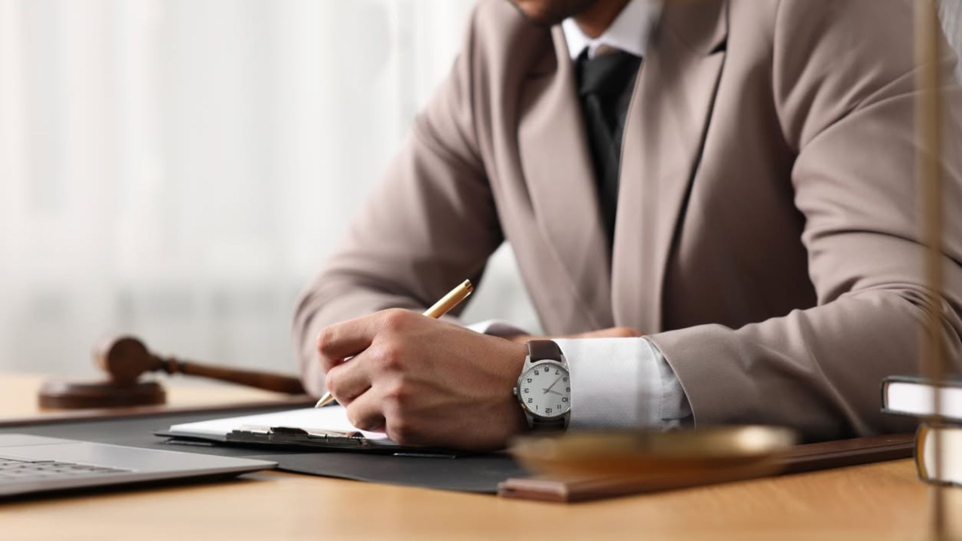 Businessperson in a beige suit writing on a clipboard at a desk, with a gavel and notebook nearby.