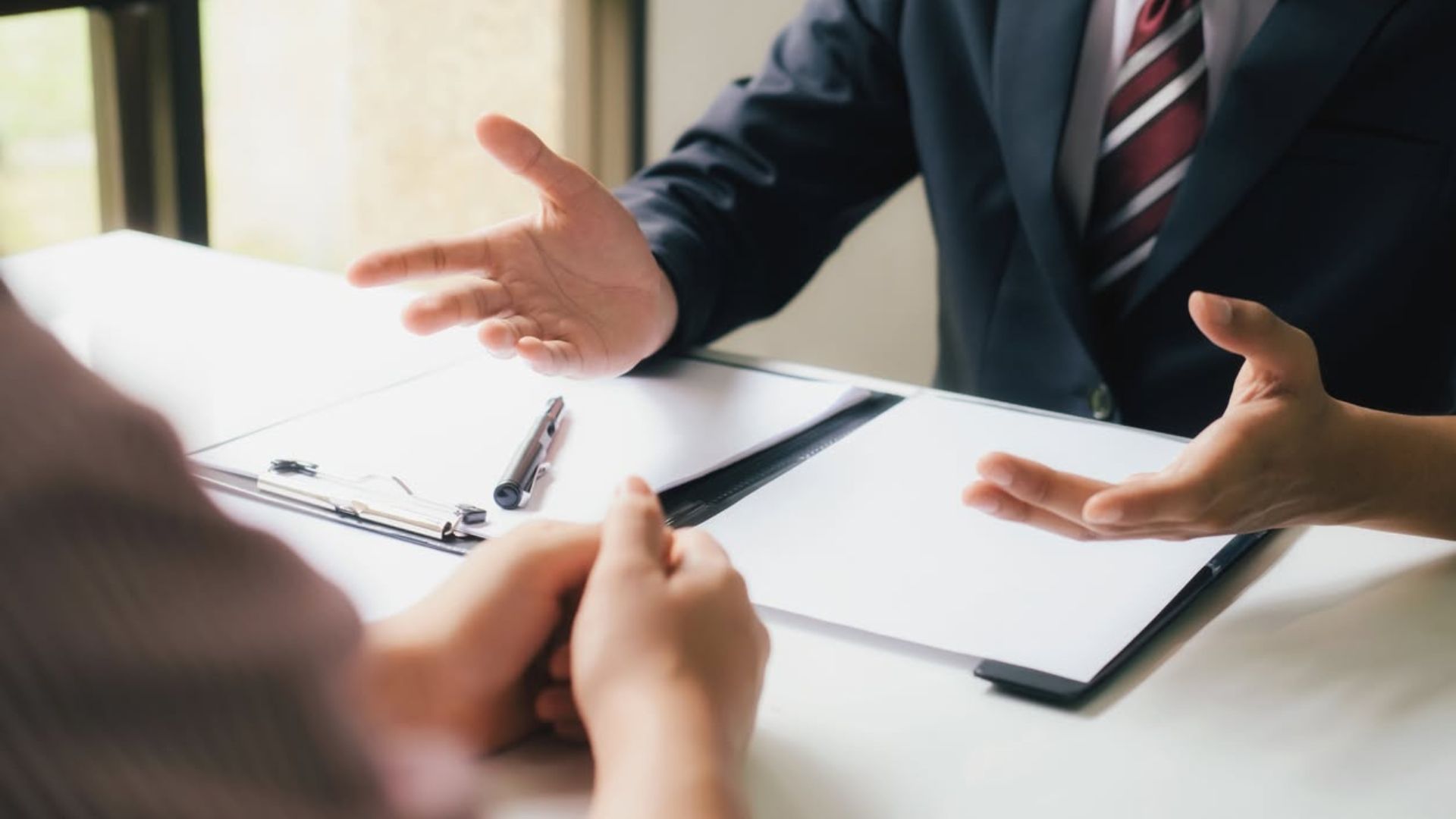 Two people in a business meeting, gesturing across a desk with documents and a pen.
