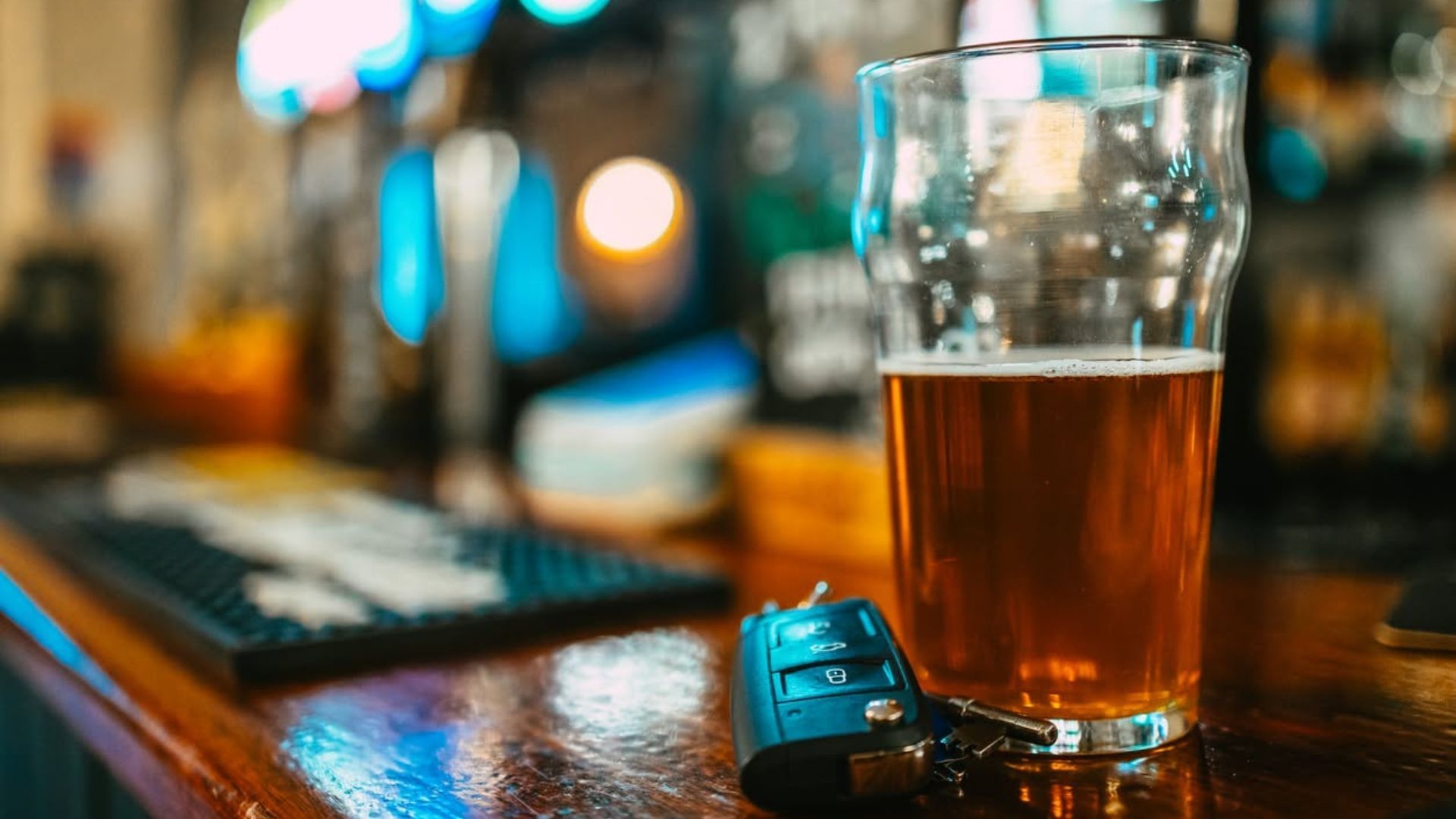 Glass of beer beside a car key on a bar counter with a blurred laptop in the background