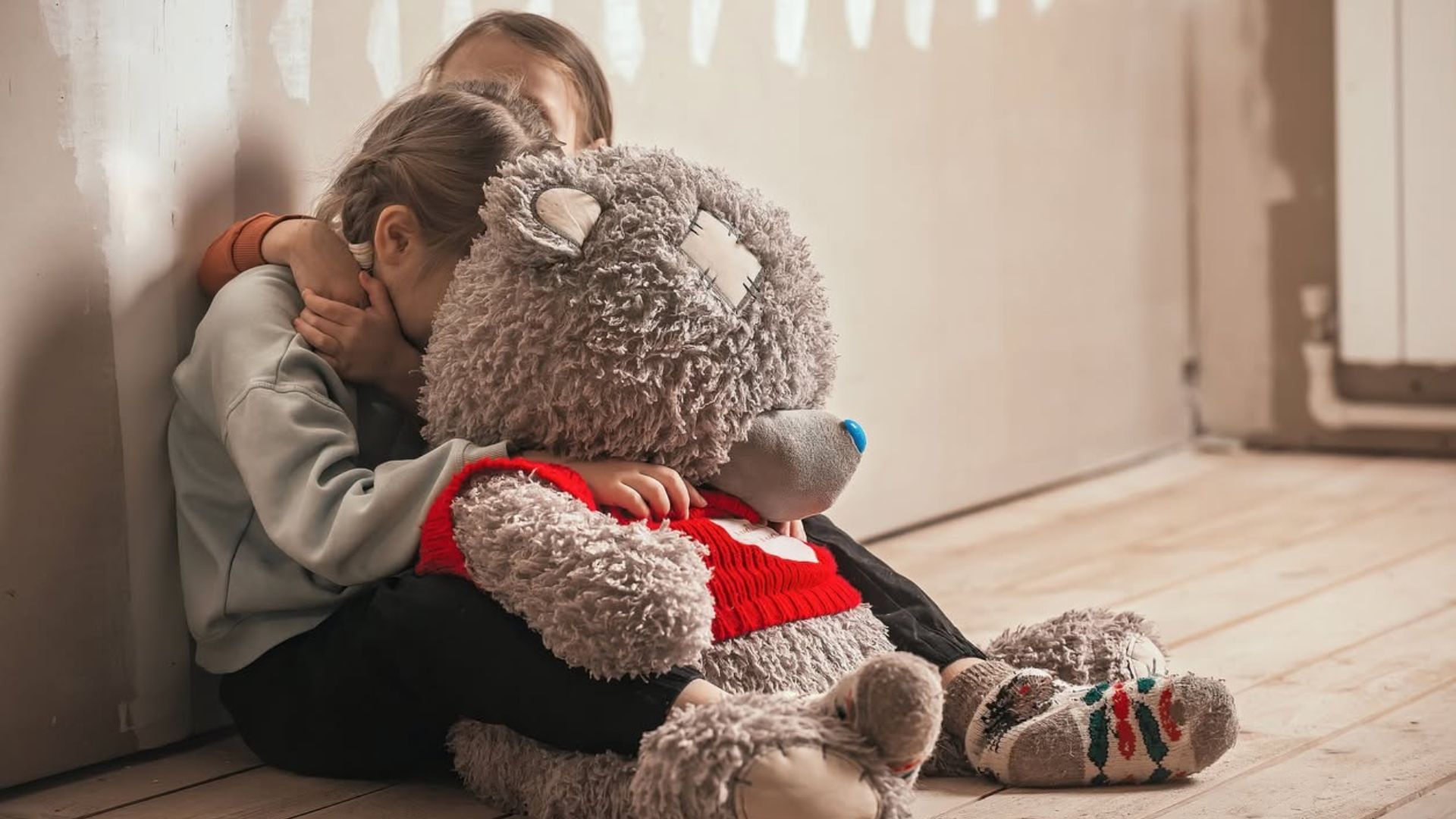 Child hugging a large gray teddy bear while sitting on the floor in warm sunlight