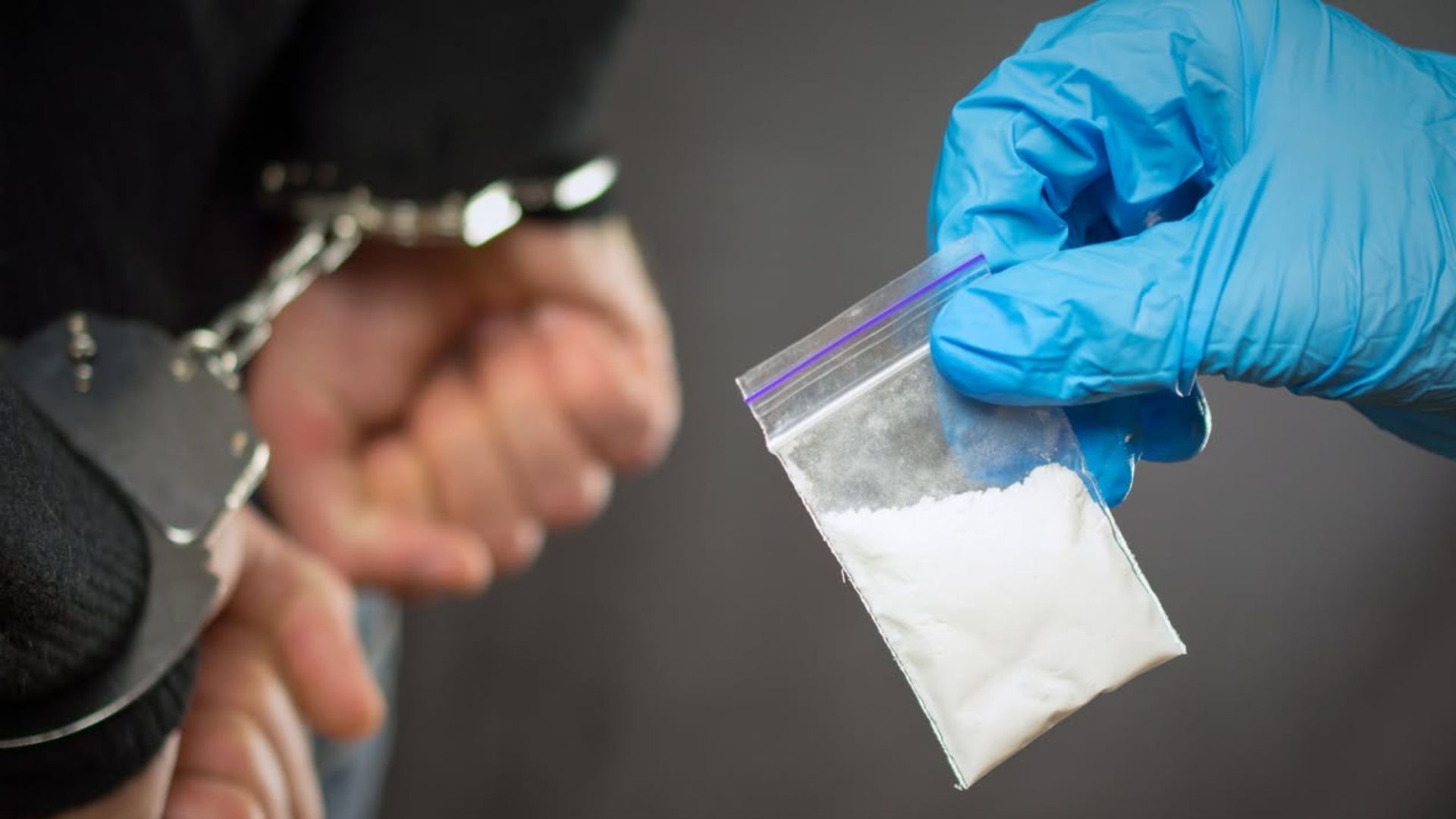 Blue-gloved hand holding a small bag of white powder beside handcuffed wrists