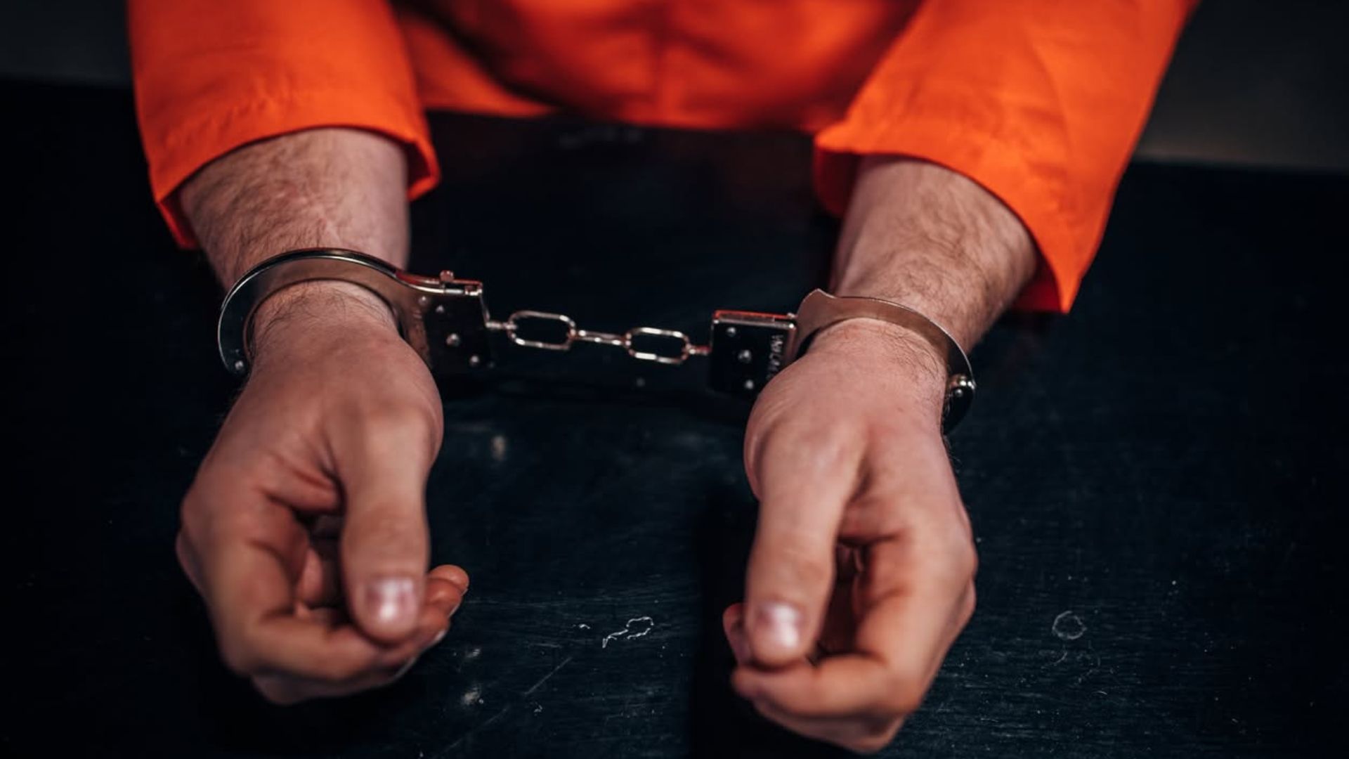 Person in orange sleeves with handcuffed wrists against a dark background
