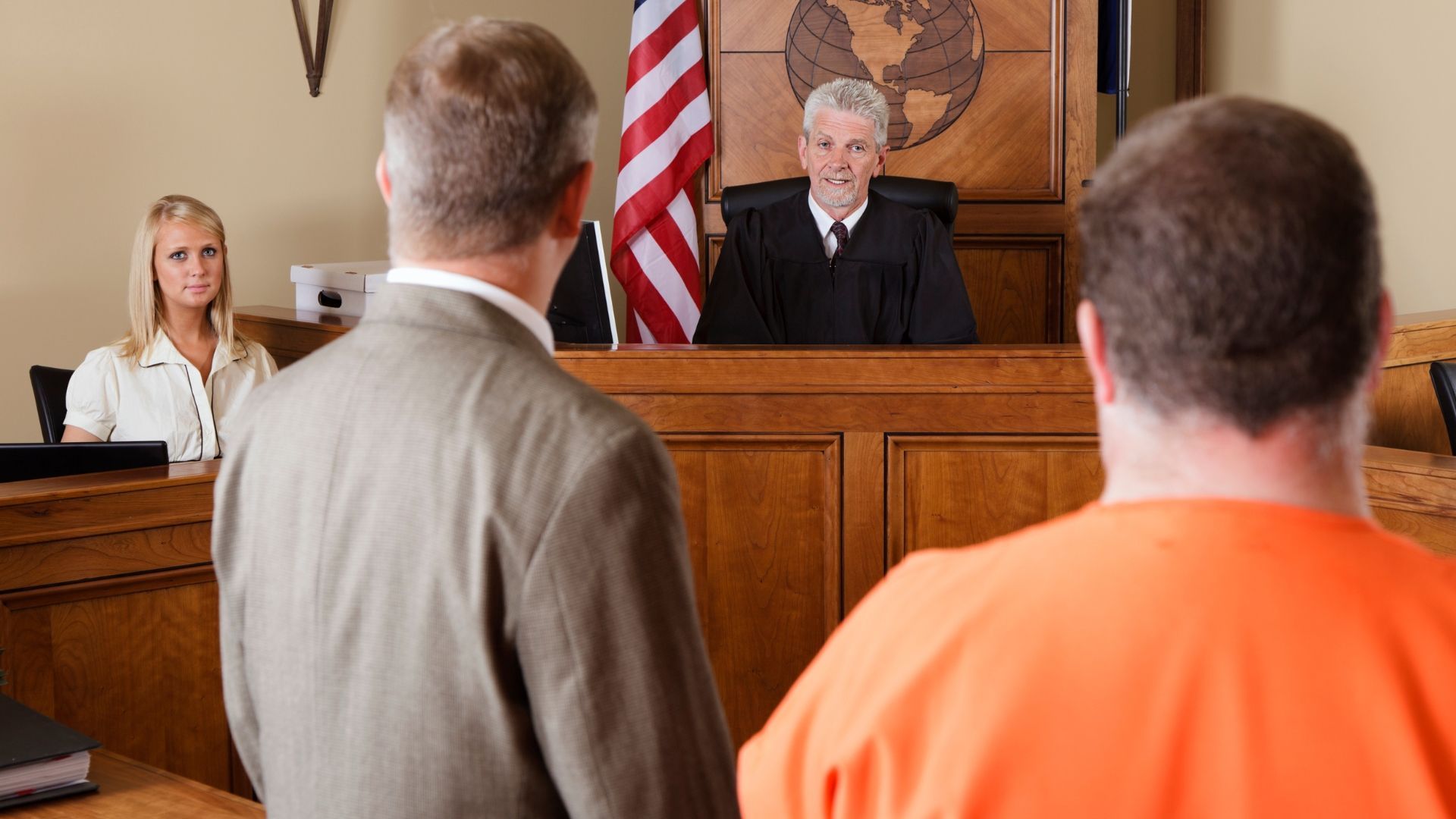 Courtroom scene with a judge at the bench and two people standing in the foreground, one in an orange shirt.