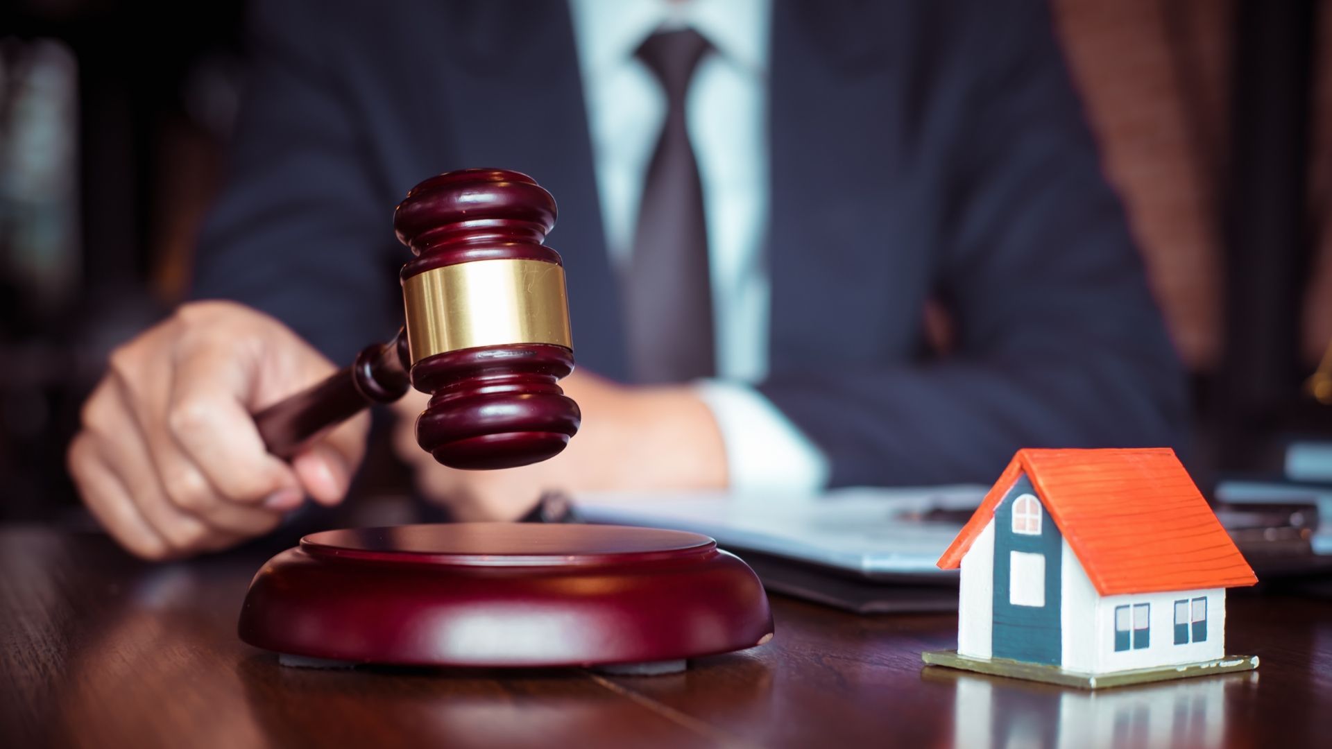 Judge’s gavel beside a small house model on a desk, with a person in a suit blurred in the background