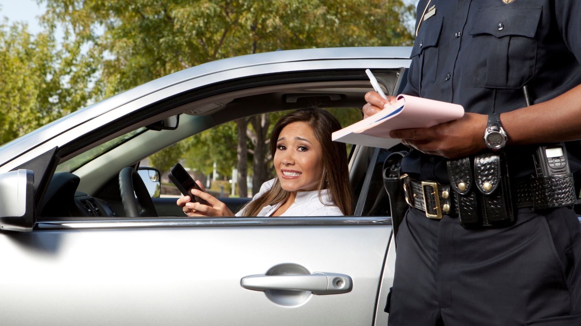 Police officer writing a ticket beside a silver car; driver smiles and holds a phone.