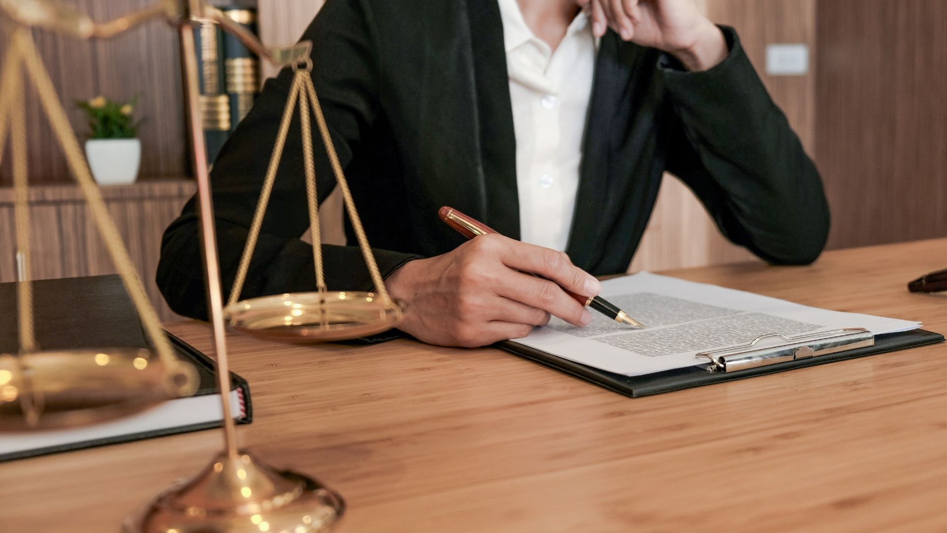 Person in a dark blazer writes in a notebook at a wooden desk with a brass scale in a legal office