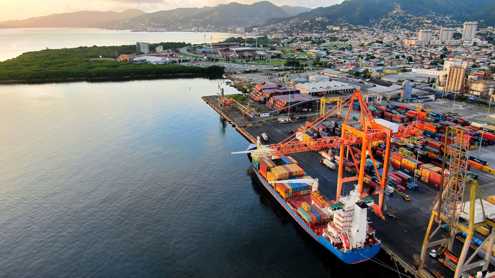 Cargo ship docked at a bustling port, cranes loading containers, city and mountains in the background.