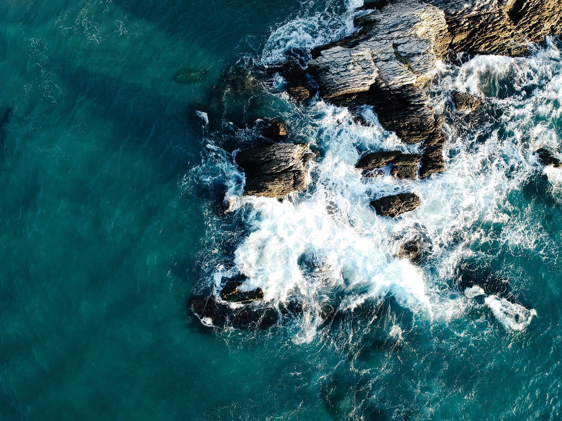Ocean waves crashing against dark, rocky shoreline; teal water.