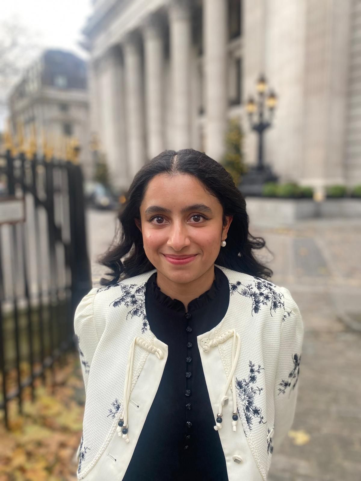 Woman with dark hair smiles outdoors; wears a black top and white jacket with floral embroidery; background is a building.