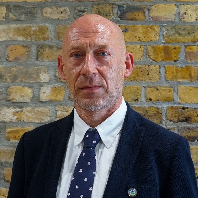 Bald man in a dark suit and patterned tie stands against a brick wall.