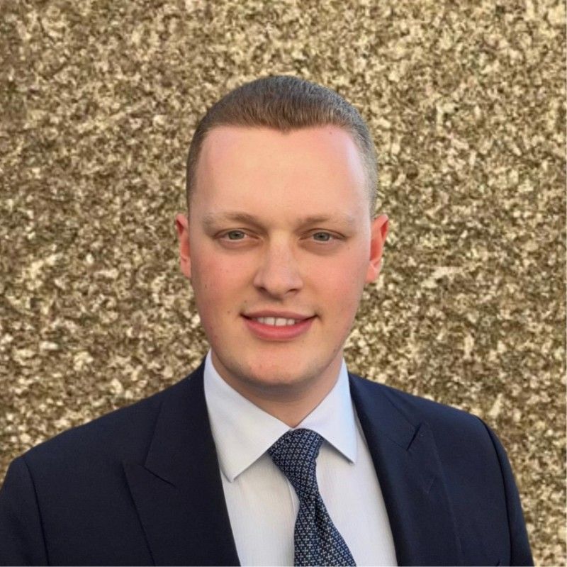 Man in a suit smiles at the camera in front of a textured, tan wall.
