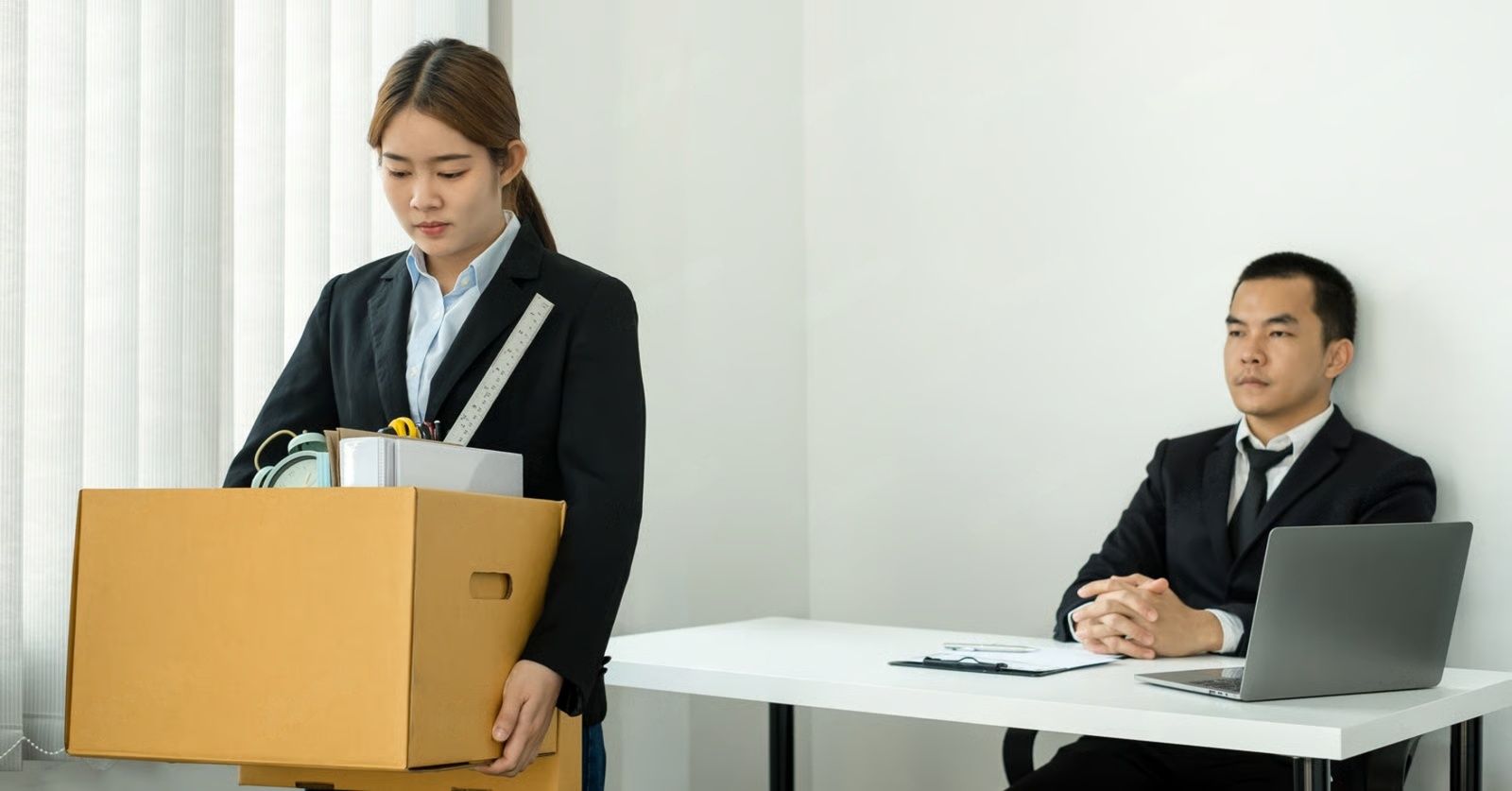 Woman carrying box of belongings, leaving office; man sits at desk.