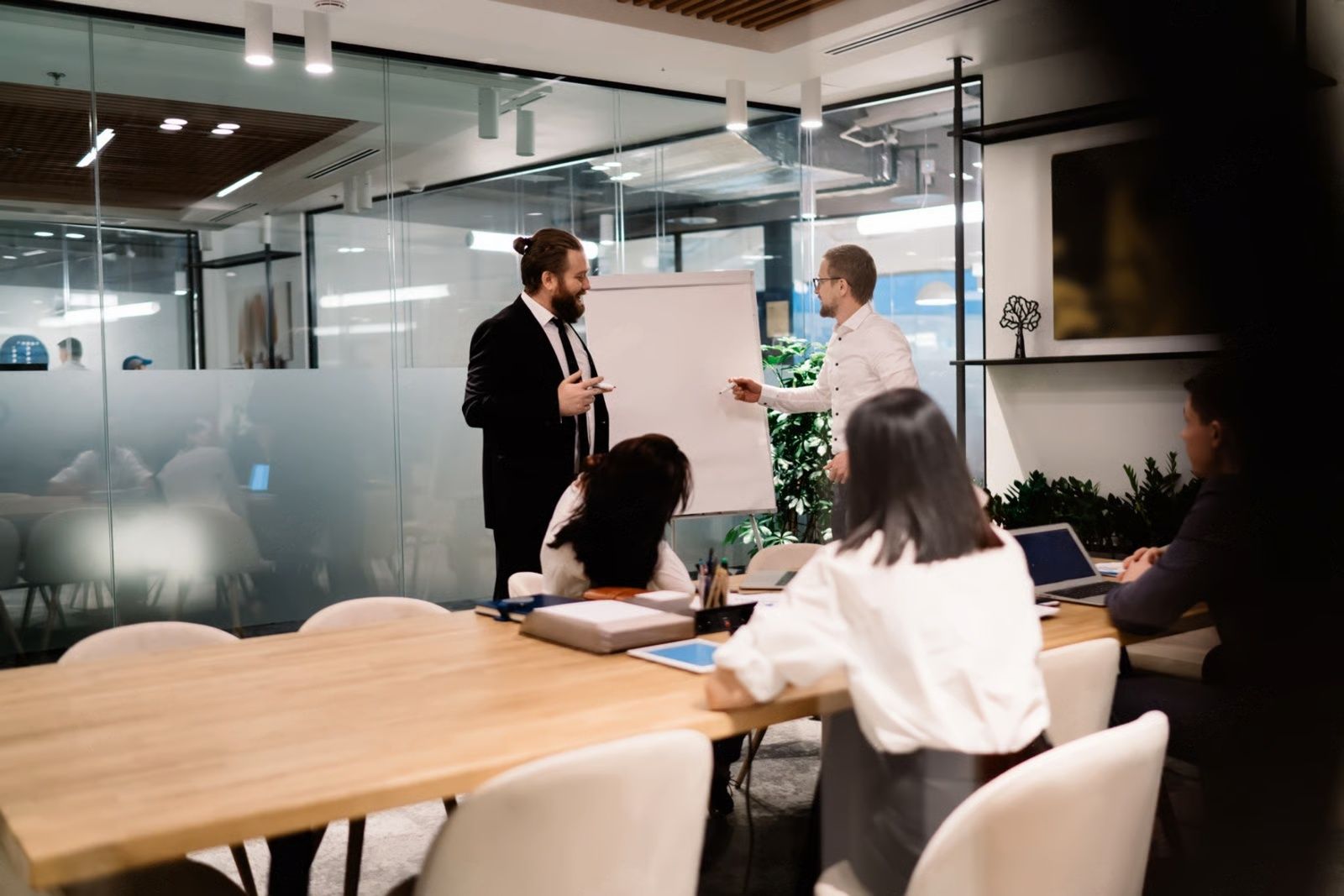 Business meeting in modern office. Two men near whiteboard, others at table with laptops.