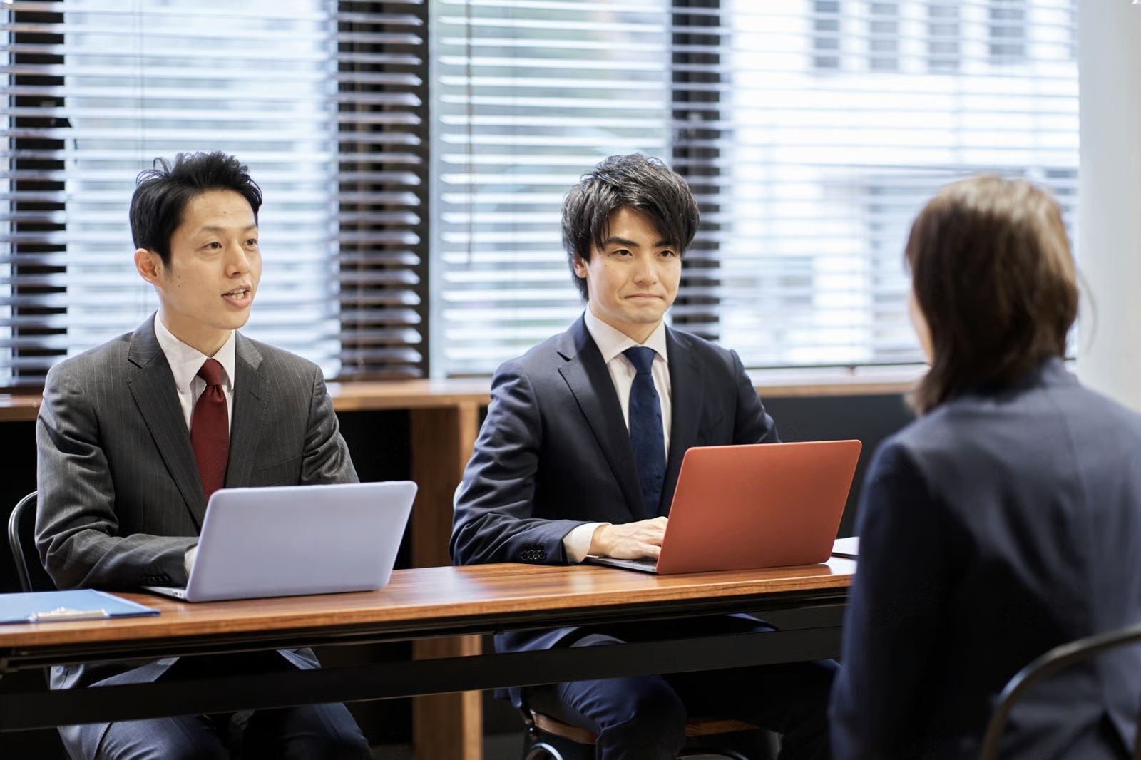 Two men in suits interview a person at a table with laptops in an office.