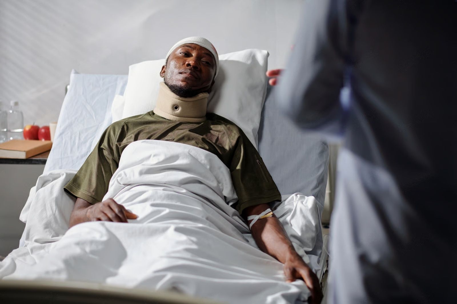 Person in hospital bed with neck brace, head bandages, and IV. Doctor at bedside.