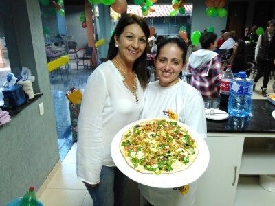 Duas mulheres sorridentes, segurando uma grande travessa de comida em uma festa realizada em ambiente fechado.