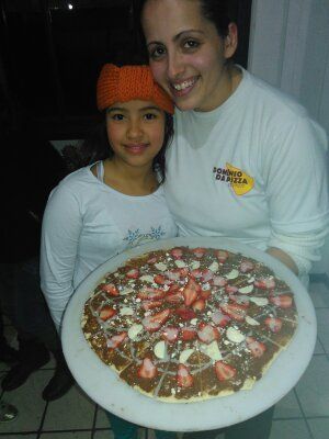 Duas pessoas sorrindo e segurando uma pizza grande em um prato em um quarto com pouca luz.