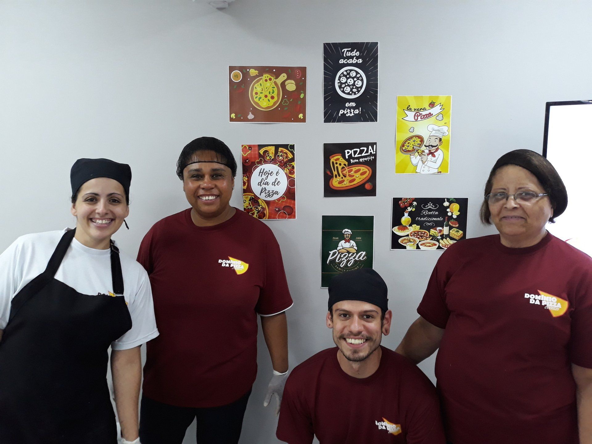 Quatro funcionários da cozinha, de avental e camisa bordô, posam sorrindo em frente a cartazes de comida na parede.
