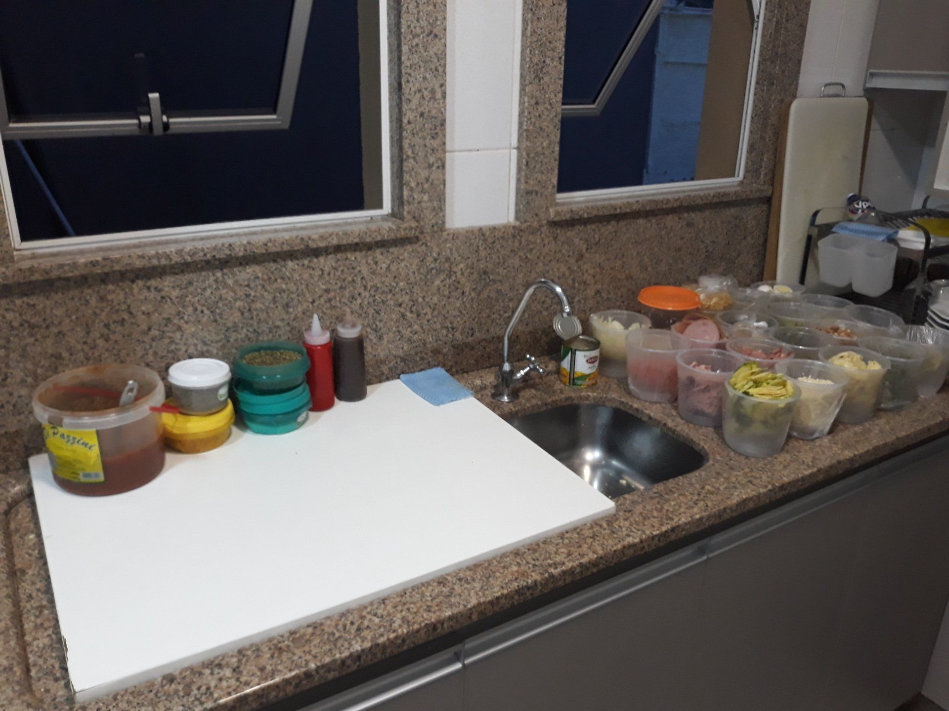 Bancada da cozinha ao lado da pia com recipientes, potes e uma tábua de corte dispostos para o preparo de alimentos.