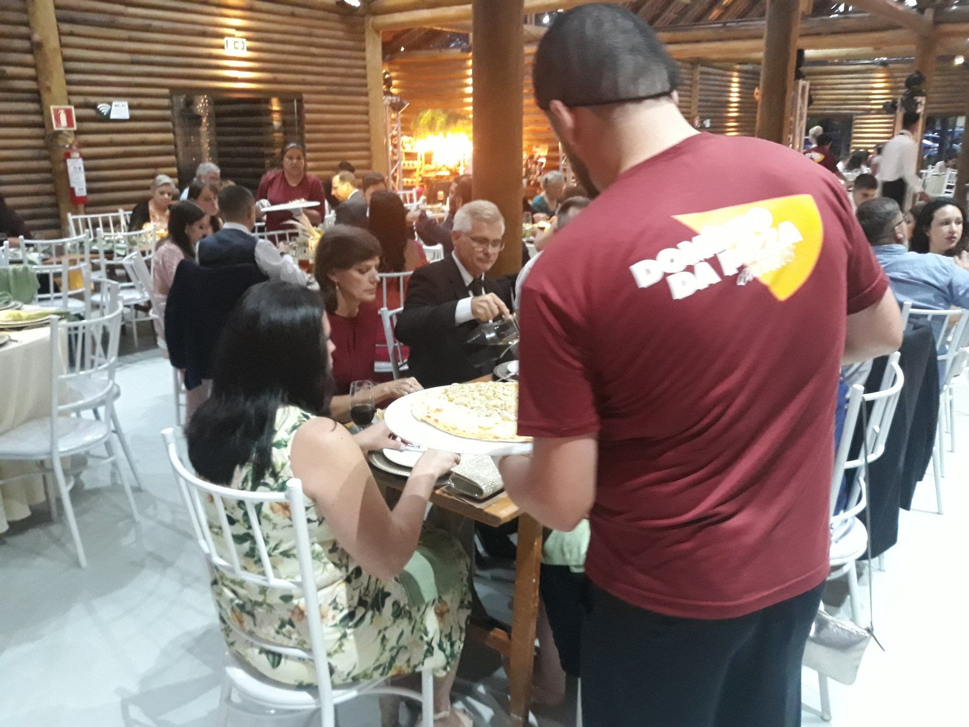 Pessoas sentadas às mesas em um restaurante rústico enquanto um garçom de camisa vermelha carrega a comida.