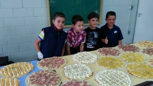 Quatro meninos estão de pé atrás de mesas cobertas com bandejas de alimentos redondos e coloridos em uma sala revestida de azulejos.