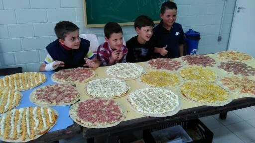 Crianças em pé atrás de mesas cobertas com várias pizzas redondas em uma sala de aula ou refeitório.