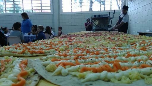 Grande mesa coberta com pizzas variadas em um refeitório iluminado, com pessoas ao fundo.