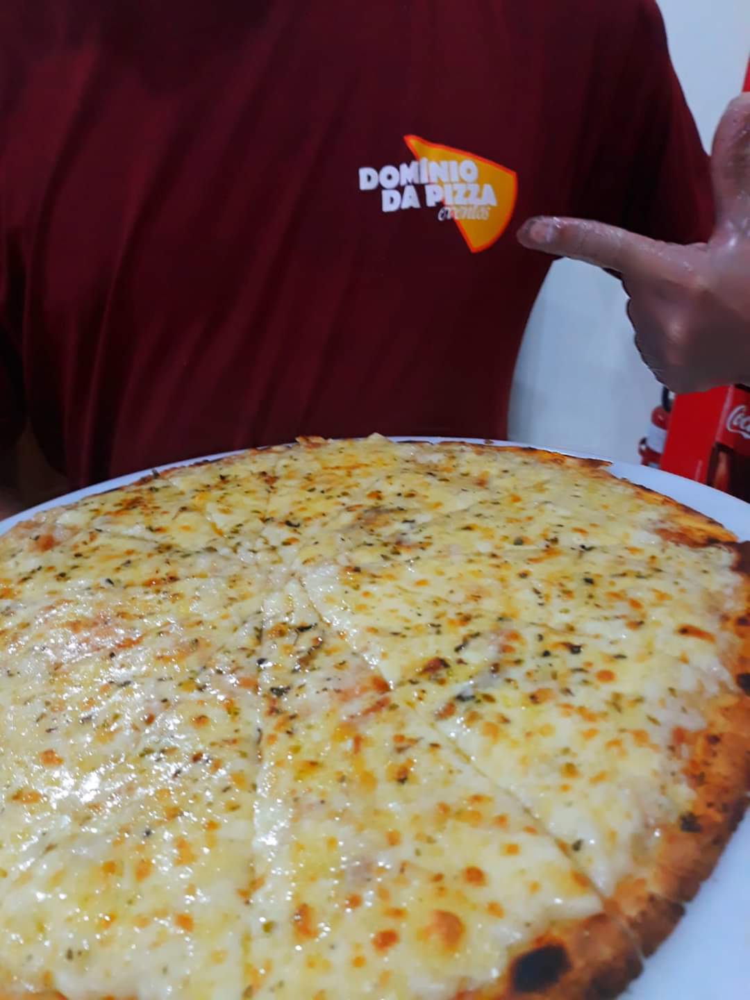 Pessoa segurando uma pizza grande de queijo em uma bandeja, vestindo uma camisa vermelha.