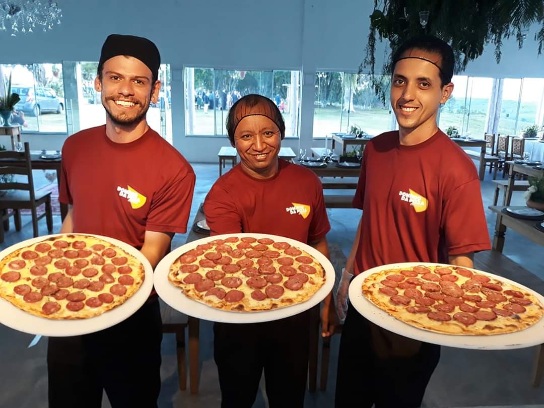 Três entregadores de pizza com camisas vermelhas segurando pizzas grandes em um restaurante.