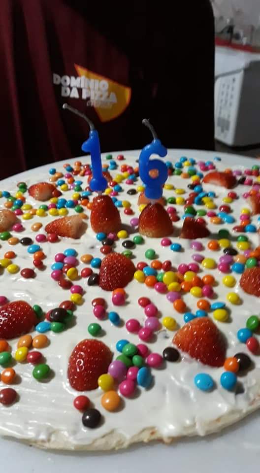 Bolo de aniversário branco coberto com confeitos coloridos e morangos, decorado com velas azuis em formato de 5 e 6.