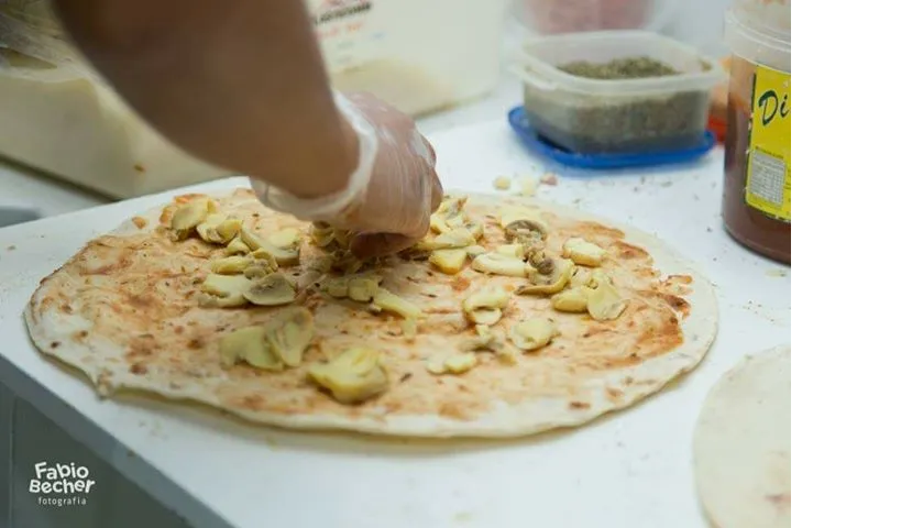 Mãos decorando uma pizza com legumes fatiados sobre uma mesa de preparo branca.