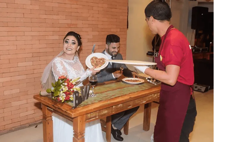 A noiva sorri para a mesa decorada enquanto um garçom de vermelho apresenta a comida ao lado do noivo.