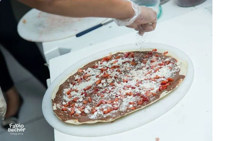 Uma pessoa polvilha queijo e tomate sobre uma pizza em uma mesa branca.